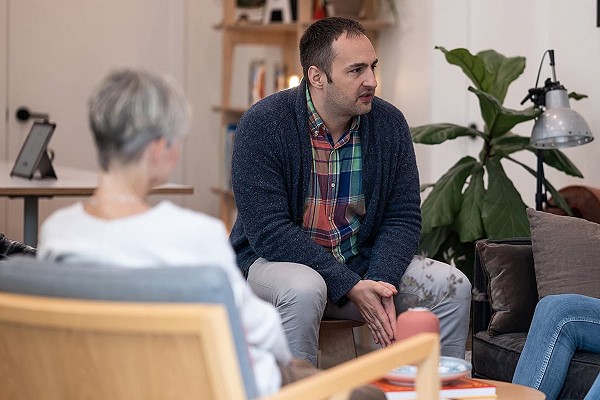 A man wearing a plaid shirt and blue sweater addresses two people in a room; his is sitting in a chair and has his hands together